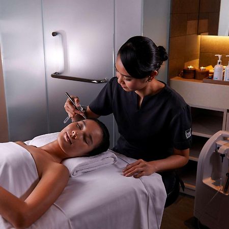 Paradox Singapore Merchant Court At Clarke Quay المظهر الخارجي الصورة The photo shows a spa or beauty therapy setting where a woman is lying on a treatment table with a towel draped over her. A professional, dressed in a dark uniform, is performing a skincare treatment on her face. Various beauty products are visible o
