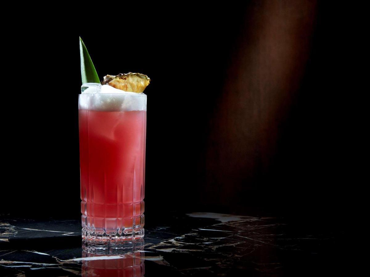 Paradox Singapore Merchant Court At Clarke Quay المظهر الخارجي الصورة The photo shows a tall cocktail glass filled with a vibrant red drink. The drink appears to have a frothy top and is garnished with a slice of pineapple and a piece of green leaf. The glass is placed on a dark, glossy surface, and the background is s
