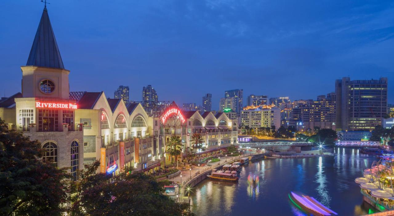 Paradox Singapore Merchant Court At Clarke Quay المظهر الخارجي الصورة The photo shows a riverside scene at dusk, featuring a waterfront area with modern buildings that have a distinctive architectural style. The structures have pointed roofs and are well-lit, creating a vibrant atmosphere. Along the river, there are bo