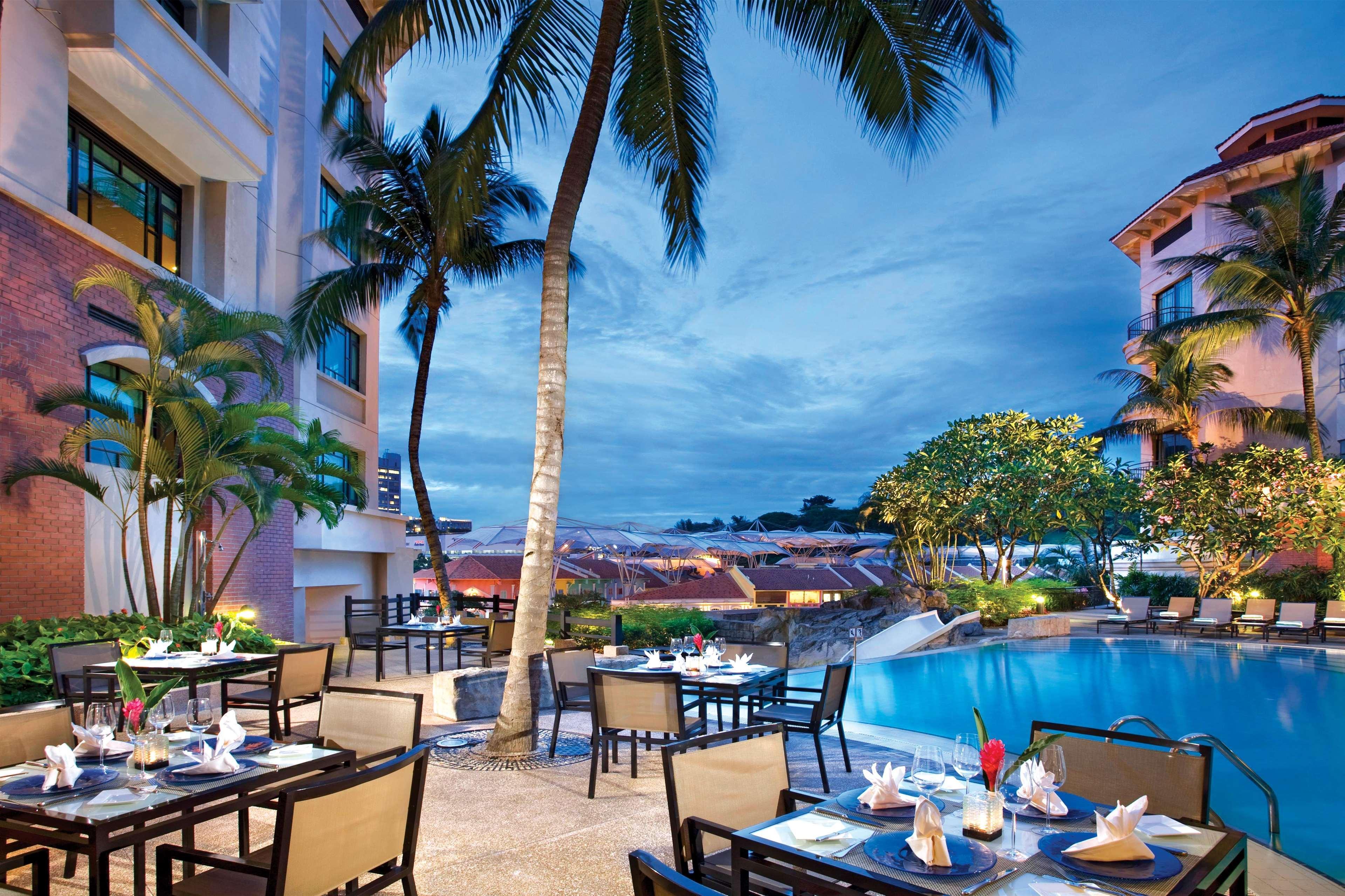 Paradox Singapore Merchant Court At Clarke Quay المطعم الصورة The photo depicts a beautiful outdoor dining area next to a shimmering pool, set against a backdrop of palm trees and a dramatic evening sky. Tables are arranged elegantly with white tablecloths, chairs, and decorative napkins, suggesting a luxurious