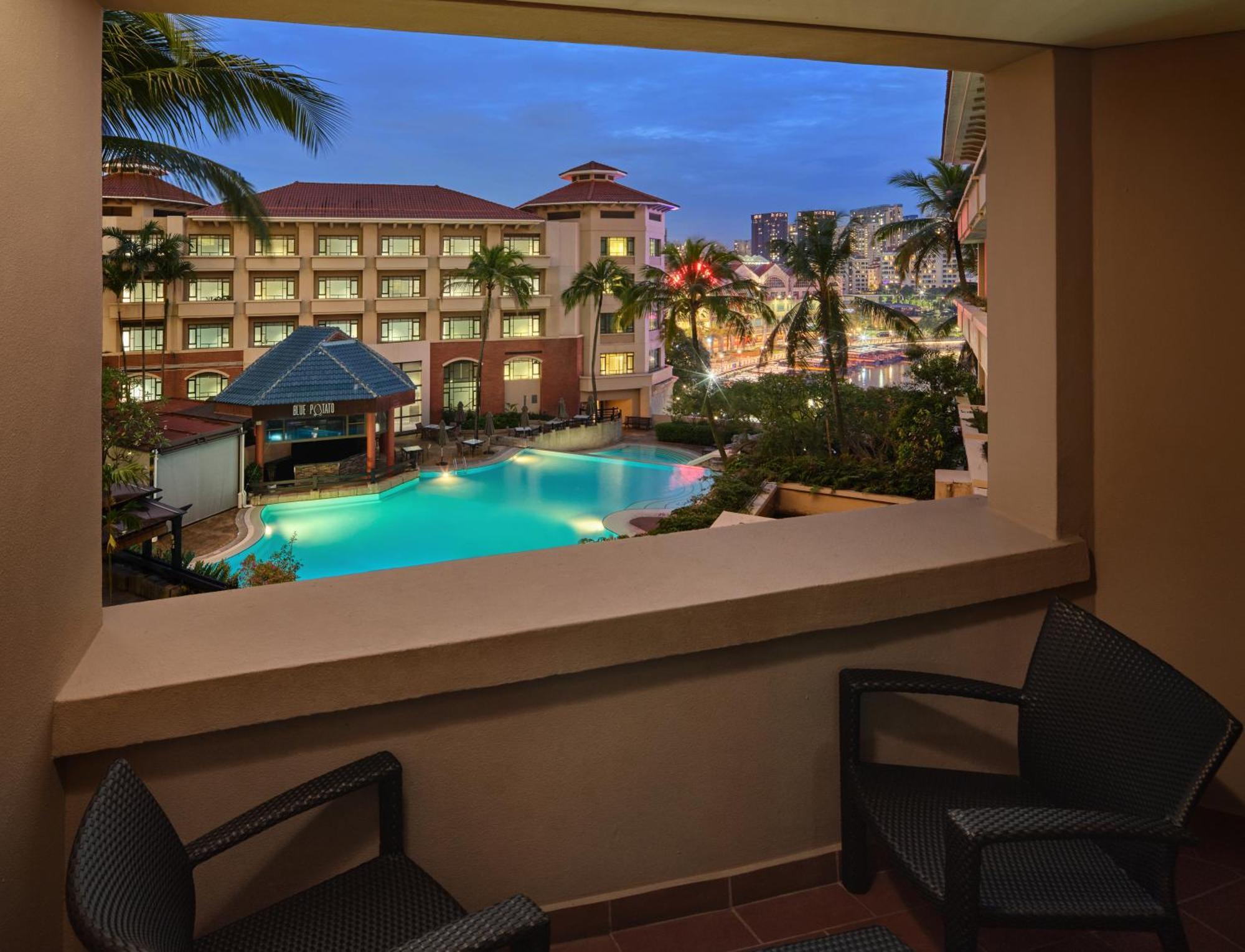 Paradox Singapore Merchant Court At Clarke Quay المظهر الخارجي الصورة The image depicts a view from a balcony overlooking a pool area of a hotel or resort. The scene features palm trees surrounding the pool, which has a blue hue, possibly illuminated by lights. In the background, there are several buildings, likely par
